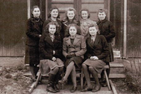 Acht Frauen werden vor einer Baracke fotografiert. Drei Frauen, darunter Nina Wassilywna Koslowa in der 1. Reihe Mitte, sitzen auf einer Bank. Die anderen fünf Frauen stehen dahinter. Die Frauen tragen Röcke oder Kleider mit Jacken oder Mänteln darüber. Einige tragen Stiefel, andere knöchelhohe Schuhe. Sie schauen in die Kamera, Vier Frauen lächeln leicht. 