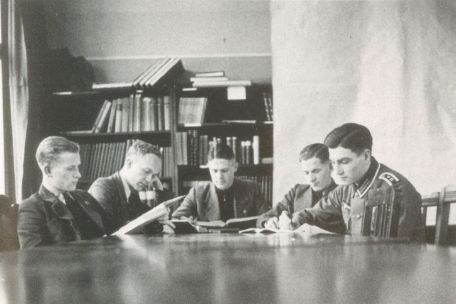 Fünf Personen sitzen an einem Tisch. Es sind Studenten der Universität. Sie haben Bücher vor sich und in den Händen. Sie schauen in die Bücher. im Hintergrund steht ein Bücherregal.